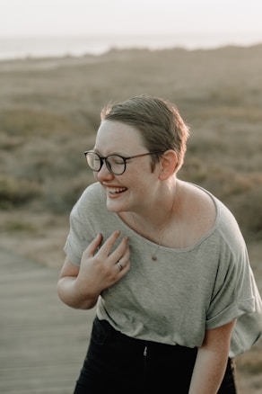 A person with short hair and glasses is laughing in an outdoor setting. The scene is lit by soft, natural light, suggesting sunset or sunrise. The person is wearing a casual gray shirt and appears relaxed and joyful. The background consists of a blurred landscape with earthy tones, possibly a field or coastal area.