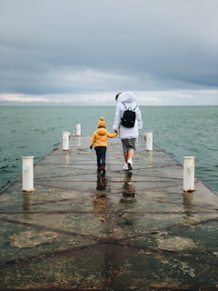 A person and a child walk hand in hand on a pier leading into a body of water. The sky is overcast, and the water is a muted green-gray color. The adult is wearing a white jacket with a hood and a black backpack, while the child is in a bright yellow jacket and a matching beanie.