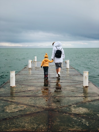 A person and a child walk hand in hand on a pier leading into a body of water. The sky is overcast, and the water is a muted green-gray color. The adult is wearing a white jacket with a hood and a black backpack, while the child is in a bright yellow jacket and a matching beanie.