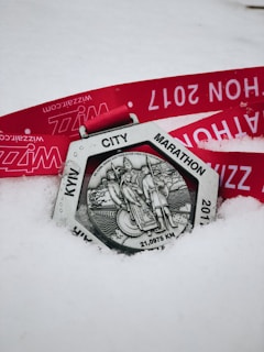 A silver medal with an intricate design featuring figures and the inscription 'KYW CITY MARATHON' and '21,0975 KM'. The medal rests on snow with a bright red ribbon that has text printed in white.