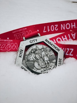 A silver medal with an intricate design featuring figures and the inscription 'KYW CITY MARATHON' and '21,0975 KM'. The medal rests on snow with a bright red ribbon that has text printed in white.