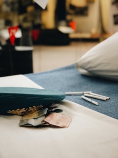 A teal military-style cap with gold embroidered leaves rests on a white cloth. Beside the cap, a few currency notes and coins are scattered. In the blurred background, a few red and black objects are visible, along with a pillow on a blue surface. White cigarettes lie nearby.