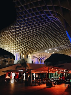 An architectural structure with a honeycomb lattice design is illuminated in a blend of warm and cool lighting. Below, an outdoor dining area with tables and chairs is set under canopies, with glowing lamps adding a cozy ambiance. Some people are seen enjoying the space.