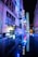 Vibrant neon signs and reflections create a cinematic atmosphere in an urban alley. Bright blue, red, and yellow lights illuminate the architecture, suggesting a lively street at night. The perspective captures both the vertical signs on the building and their mirrored reflection on a glossy surface below.