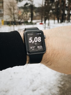 A person is wearing a smartwatch on their wrist, displaying a distance of 5.08 kilometers. The background is a snowy outdoor setting with some trees and buildings visible.