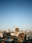 A cityscape featuring a prominent telecommunications tower standing tall amid various modern and historic buildings. The foreground includes a river with a bridge and lush greenery. The sky is expansive and clear, enhancing the architectural features and urban layout.