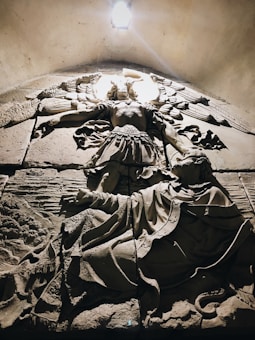A detailed stone relief features an angelic figure with wide wings, raised arms, and ornate garments, positioned above a reclining, bearded figure. The scene is dimly lit, with light highlighting the angel from above, casting shadows that emphasize the intricate textures of the carving.