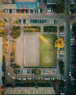 An aerial view of a city block featuring a sports area with a basketball court and a tennis court. Surrounding the courts are parked cars along the streets and lush greenery with trees providing shade. Buildings frame the scene, and sunlight casts long shadows over the ground.