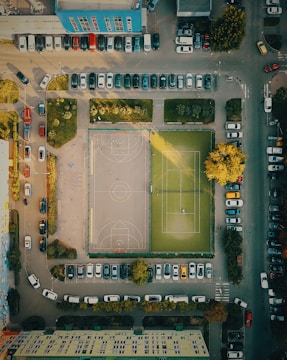 An aerial view of a city block featuring a sports area with a basketball court and a tennis court. Surrounding the courts are parked cars along the streets and lush greenery with trees providing shade. Buildings frame the scene, and sunlight casts long shadows over the ground.