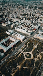 Aerial view of an urban landscape with densely packed buildings and streets forming a grid pattern. There is a large circular park area with trees and pathways situated prominently, surrounded by roads and vehicles.