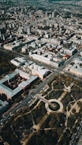 Aerial view of an urban landscape with densely packed buildings and streets forming a grid pattern. There is a large circular park area with trees and pathways situated prominently, surrounded by roads and vehicles.