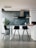 Minimalist kitchen interior with a modern design, featuring a gray concrete wall, a white countertop with black bar stools, and a potted plant in the corner. Various items are arranged on the countertop including jars and a water bottle.