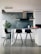 Minimalist kitchen interior with a modern design, featuring a gray concrete wall, a white countertop with black bar stools, and a potted plant in the corner. Various items are arranged on the countertop including jars and a water bottle.
