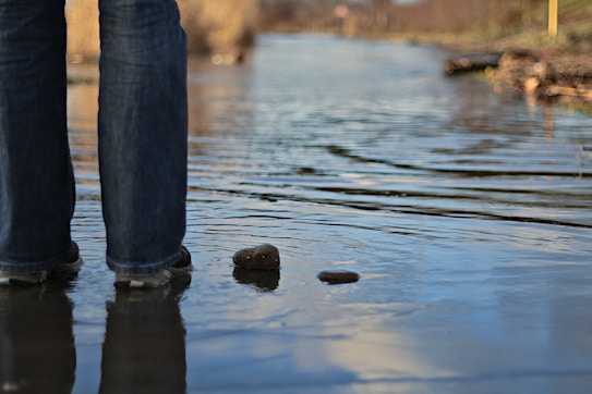 A pair of legs in jeans stands in shallow water on a flooded path, creating ripples. The surrounding area shows blurred trees and ground, indicating a natural setting.