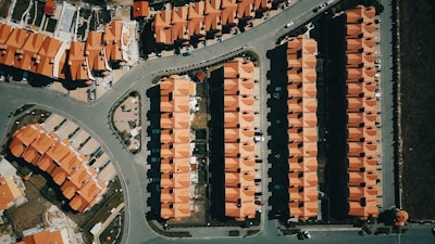 Aerial view of a suburban area with rows of houses featuring uniform red roofs. The layout is highly organized with each house aligned neatly in rows. Roads and pathways are visible, dividing the neighborhood into sections. Cars are parked in driveways and along the streets.