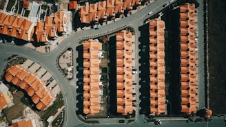 Aerial view of a suburban area with rows of houses featuring uniform red roofs. The layout is highly organized with each house aligned neatly in rows. Roads and pathways are visible, dividing the neighborhood into sections. Cars are parked in driveways and along the streets.