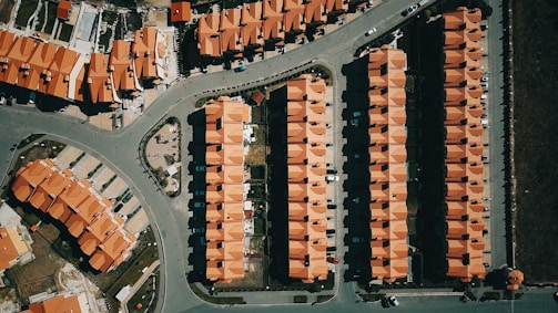 Aerial view of a suburban area with rows of houses featuring uniform red roofs. The layout is highly organized with each house aligned neatly in rows. Roads and pathways are visible, dividing the neighborhood into sections. Cars are parked in driveways and along the streets.