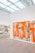 A contemporary art gallery features a large panel with abstract human figures set against a vivid orange background. The artwork incorporates mixed media, including photographic elements and geometric patterns. The gallery space is well-lit with natural light filtering through a glass ceiling, and the floor is made of light wood.