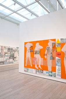A contemporary art gallery features a large panel with abstract human figures set against a vivid orange background. The artwork incorporates mixed media, including photographic elements and geometric patterns. The gallery space is well-lit with natural light filtering through a glass ceiling, and the floor is made of light wood.