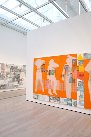 A contemporary art gallery features a large panel with abstract human figures set against a vivid orange background. The artwork incorporates mixed media, including photographic elements and geometric patterns. The gallery space is well-lit with natural light filtering through a glass ceiling, and the floor is made of light wood.
