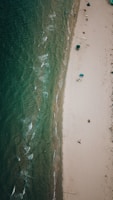 An aerial view of a coastline showcases a sandy beach parallel to a body of greenish-blue water with gentle waves. Sparse beachgoers are scattered along the shore, some with umbrellas providing shade.