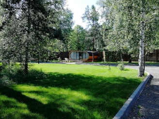 A lush green lawn surrounded by tall trees with a unique cylindrical structure featuring large windows and an orange base positioned in the background. The scene is peaceful and well-maintained, with a clear pathway curving through the greenery.
