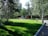 A lush green lawn surrounded by tall trees with a unique cylindrical structure featuring large windows and an orange base positioned in the background. The scene is peaceful and well-maintained, with a clear pathway curving through the greenery.
