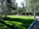 A lush green lawn surrounded by tall trees with a unique cylindrical structure featuring large windows and an orange base positioned in the background. The scene is peaceful and well-maintained, with a clear pathway curving through the greenery.