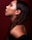Profile view of a woman with smooth skin and long brown hair, featuring subtle makeup with reddish eyeshadow. She is wearing small hoop earrings and positioned against a deep red background.