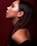Profile view of a woman with smooth skin and long brown hair, featuring subtle makeup with reddish eyeshadow. She is wearing small hoop earrings and positioned against a deep red background.