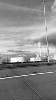 An industrial scene with large storage tanks and smokestacks emitting smoke in a factory area. The foreground includes a road with visible lanes and markings. There's a tall streetlight to the right, and the sky is filled with clouds, giving a moody and overcast appearance.