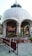 A large, white dome structure with decorative arches and intricate carvings at the top. It is supported by white pillars, and red potted plants line its perimeter. A barricade with Hindi text stands in the foreground, and people can be seen inside the pavilion.