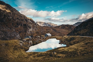 A serene mountain landscape features a small lake reflecting the blue sky and scattered clouds. Two cabins are nestled among rocky hills and grassy terrain, creating a peaceful and secluded setting.