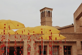 Multiple bright yellow umbrellas are open in an outdoor setting with a brick building backdrop. The umbrellas are adorned with red rectangular embellishments, and part of a traditional architecture is visible, featuring a wind tower.