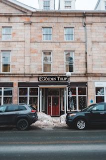 A multi-story brick building with large rectangular windows. The central focus is a storefront called 'The Golden Tulip' with a prominent black and gold sign. Two black cars are parked in front on the street, and there are snow piles at the sidewalk's edge. The building has a classic, historic look with stone detailing around the windows and doors.