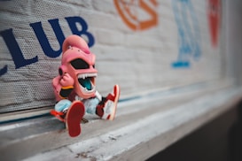 A cartoonish figurine depicting a character with a large open mouth and exaggerated features is sitting against a wall. The character is pink and wearing bright red shoes, set against a background with graffiti-style lettering and symbols.