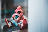 A brightly colored cartoonish figurine with a pink alien-like face and a wide open mouth revealing large white teeth. The figure is wearing sunglasses and sports a trendy outfit including sneakers with text on them. It is seated in a casual position, suggesting a playful or relaxed vibe. The background appears blurred, emphasizing the character.