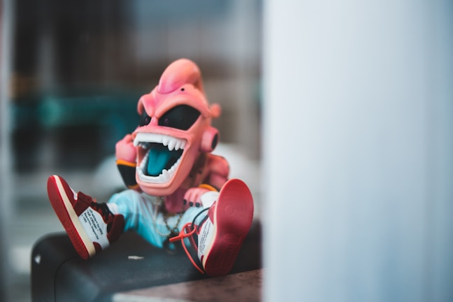 A brightly colored cartoonish figurine with a pink alien-like face and a wide open mouth revealing large white teeth. The figure is wearing sunglasses and sports a trendy outfit including sneakers with text on them. It is seated in a casual position, suggesting a playful or relaxed vibe. The background appears blurred, emphasizing the character.