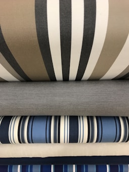 Several rolls of fabric are stacked upon one another, each with different patterns. The top roll features wide, vertical stripes in shades of beige and black. Below it is a roll with a smooth, plain gray texture. The third roll displays narrow, vertical stripes in blue and white. The bottom roll alternates between dark and light blue with thin, vertical stripes.