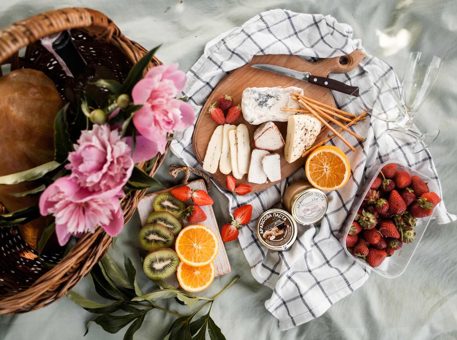 From above: a round board of three cheeses with grissini, sliced oranges, kiwi, a tray of strawberries and pink peonies on a grey-green blanket