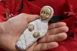 A hand gently cradles a small, intricately detailed figurine of a baby in swaddling clothes. The background is filled with vibrant red poinsettia flowers, adding a festive touch.
