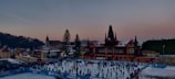 A bustling ice skating rink filled with people engaged in skating activities. The rink is surrounded by blue barriers displaying text, while a historic building with a tower and pointed roof stands prominently in the background. The setting is urban with a mix of traditional architecture and a clear evening sky transitioning from blue to pink.