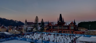A bustling ice skating rink filled with people engaged in skating activities. The rink is surrounded by blue barriers displaying text, while a historic building with a tower and pointed roof stands prominently in the background. The setting is urban with a mix of traditional architecture and a clear evening sky transitioning from blue to pink.