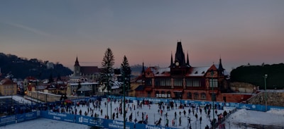 A bustling ice skating rink filled with people engaged in skating activities. The rink is surrounded by blue barriers displaying text, while a historic building with a tower and pointed roof stands prominently in the background. The setting is urban with a mix of traditional architecture and a clear evening sky transitioning from blue to pink.