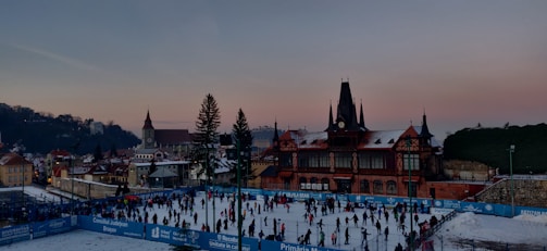 A bustling ice skating rink filled with people engaged in skating activities. The rink is surrounded by blue barriers displaying text, while a historic building with a tower and pointed roof stands prominently in the background. The setting is urban with a mix of traditional architecture and a clear evening sky transitioning from blue to pink.