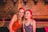 Two young women are smiling and posing for a photo in a warmly lit venue. They are standing close to each other in front of a wooden lattice wall decorated with flags bearing the word 'LAMBS' and the phrase 'WE'VE GOT History'. One woman has blonde hair and is wearing a brown sleeveless top, while the other has red hair and is wearing a purple sleeveless top. A drink with a straw is visible on the table behind them.