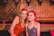Two young women are smiling and posing for a photo in a warmly lit venue. They are standing close to each other in front of a wooden lattice wall decorated with flags bearing the word 'LAMBS' and the phrase 'WE'VE GOT History'. One woman has blonde hair and is wearing a brown sleeveless top, while the other has red hair and is wearing a purple sleeveless top. A drink with a straw is visible on the table behind them.