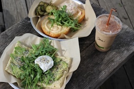 A wooden table adorned with two plates of food on brown parchment paper. One plate holds avocado toast topped with arugula, a poached egg, and seasoning. The other features a sandwich with arugula and a creamy sauce, accompanied by pickles. A cup labeled 'Chai' filled with an iced beverage and a straw is placed next to the plates.