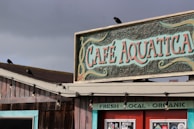 A rustic café with a wooden exterior and a vintage-style sign reading 'Café Aquatica' in bold, artistic lettering. The roof features a few birds perched atop. The weathered wood and hanging lights contribute to a cozy, coastal atmosphere. Additional signage below indicates offerings of fresh, local, and organic products.