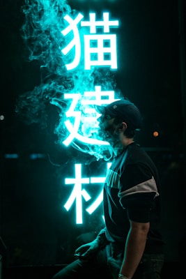 A person stands next to a glowing neon sign with stylized Asian characters. The neon emits a vibrant blue light, illuminating smoke that swirls around the person. The scene has a moody atmosphere with dark shadows contrasting against the bright lettering.